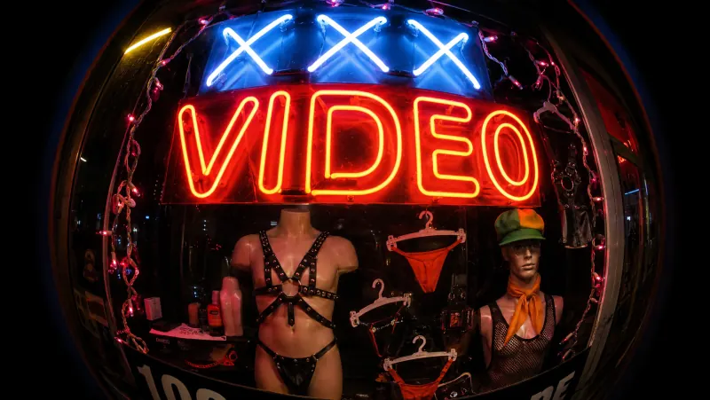 Neon-lit sex shop window at night with XXX video signage, lingerie and mannequins, evoking compulsive sexual behaviour